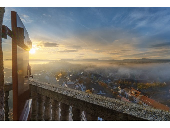 Blick von einem Balkon auf eine nebelverhangene Stadt im Morgenlicht. Die Sonne geht am Horizont auf und taucht die Szene in warmes Licht., &copy; Stadt N&uuml;rtingen