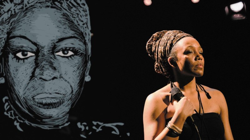 Eine Frau mit Mikrofon steht auf einer B&uuml;hne vor einem gro&szlig;en Portr&auml;t von Nina Simone. Sie tr&auml;gt ein schwarzes Kleid und ein Kopftuch.