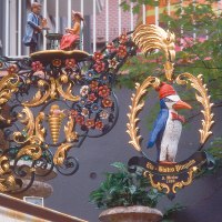 Ein kunstvoll verziertes Schild mit einem Pinguin und Figuren, umgeben von Blumenornamenten, hängt an einer Wand am Eugensplatz., © Stuttgart-Marketing GmbH