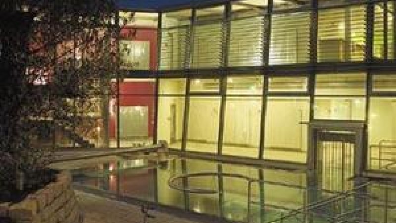 Modernes Gebäude mit Glasfassade, beleuchtet bei Nacht. Ein kleiner Pool oder Wasserfläche spiegelt das Licht wider. Bäume im Vordergrund., © Schwäbische Alb