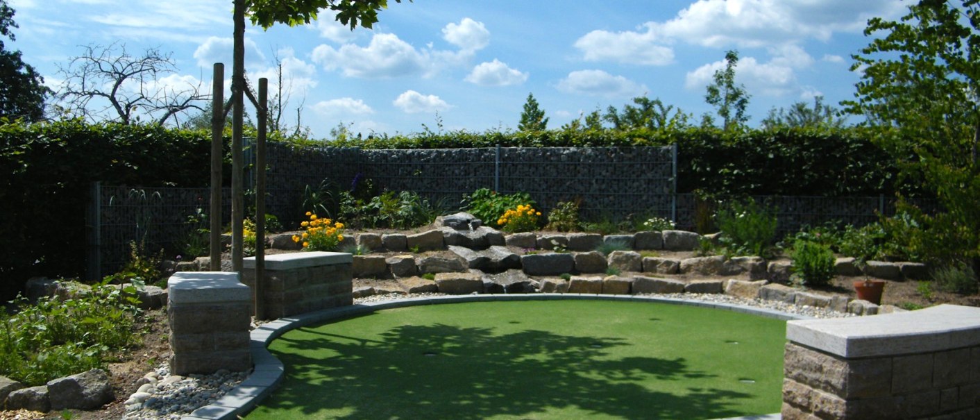 Minigolfplatz mit künstlichem Grün, umgeben von Pflanzen und Steinmauern. Ein Baum spendet Schatten, der Himmel ist blau mit weißen Wolken., © Anja Beyerle & Ralf Bähr GbR