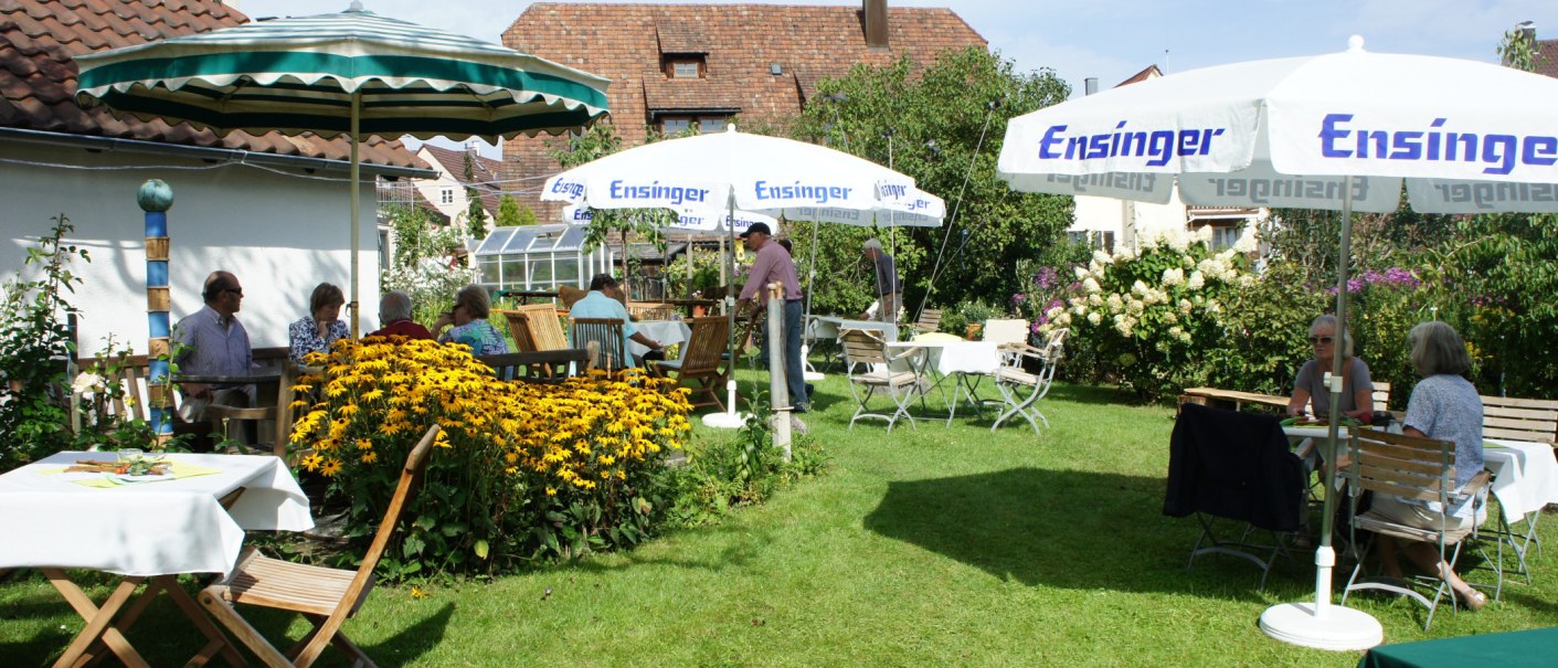 Ein sommerlicher Garten mit Menschen, die unter Sonnenschirmen an Tischen sitzen, umgeben von blühenden Blumen und grüner Vegetation., © Weinbau Knobloch-Wolfrum Ein sommerlicher Garten mit Menschen, die unter Sonnenschirmen an Tischen sitzen, umgeben von blühenden Blumen und grüner Vegetation., © Weinbau Knobloch-Wolfrum