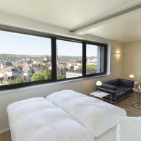 Modernes Apartment mit großem Fenster, Blick auf die Stadt, Doppelbett, schwarzem Sofa und kleinem Tisch mit Lampe., © Büroma-Apart Apartmentvermietung GmbH
