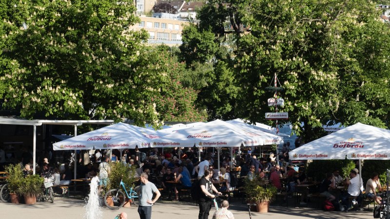 Biergarten im Schlossgarten mit vielen Menschen unter gro&szlig;en Sonnenschirmen. Ein Brunnen im Vordergrund, umgeben von gr&uuml;nen B&auml;umen und Geb&auml;uden im Hintergrund., &copy; Michele Scognamillo