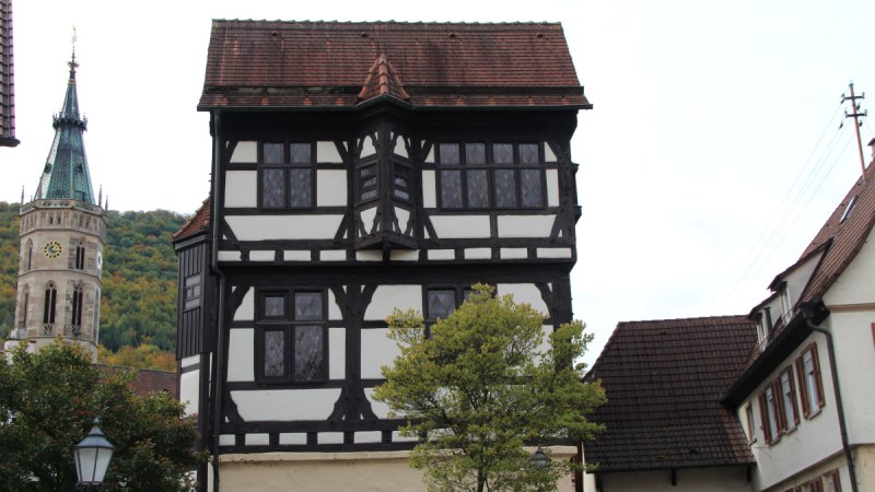 Ein Fachwerkhaus mit markantem Erker, daneben ein Kirchturm. Im Vordergrund ein Baum und eine Straßenlaterne.
