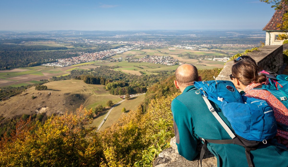 Zwei Personen mit Rucksäcken blicken von der Burg Teck auf eine weite Landschaft mit Feldern und Dörfern unter blauem Himmel., © hochgehberge