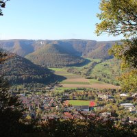 Ausblick vom Nägelesfelsen auf eine Stadt mit Sportplatz, umgeben von grünen Hügeln und Wäldern unter klarem Himmel., © Bad Urach Tourismus