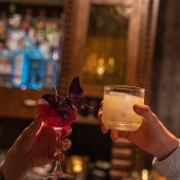 Zwei Männer stoßen in einer Bar mit Cocktails an, einer in einem karierten Sakko, der andere in einem hellen Hemd. Im Hintergrund sind Flaschen zu sehen., © SMG, Martina Denker Zwei Männer stoßen in einer Bar mit Cocktails an, einer in einem karierten Sakko, der andere in einem hellen Hemd. Im Hintergrund sind Flaschen zu sehen., © SMG, Martina Denker