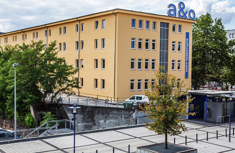 Gelbes Gebäude mit a&o Hostel Logo, umgeben von Bäumen und einem gepflasterten Platz. Im Vordergrund ein Baum und Laternen., © A&O Hostels Marketing GmbH Gelbes Gebäude mit a&o Hostel Logo, umgeben von Bäumen und einem gepflasterten Platz. Im Vordergrund ein Baum und Laternen., © A&O Hostels Marketing GmbH