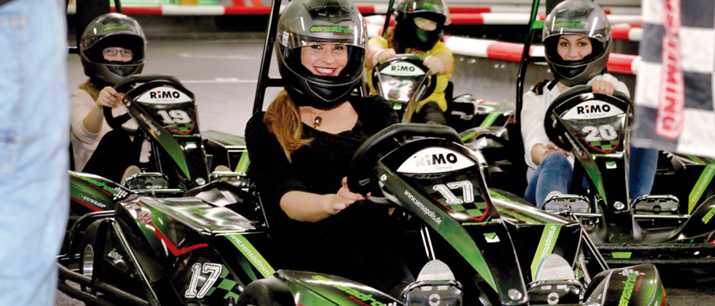 Vier Personen fahren in Karts auf einer Indoor-Kartbahn. Sie tragen Helme und lächeln. Die Karts sind schwarz-grün und nummeriert., © Sensapolis Sindelfingen Vier Personen fahren in Karts auf einer Indoor-Kartbahn. Sie tragen Helme und lächeln. Die Karts sind schwarz-grün und nummeriert., © Sensapolis Sindelfingen
