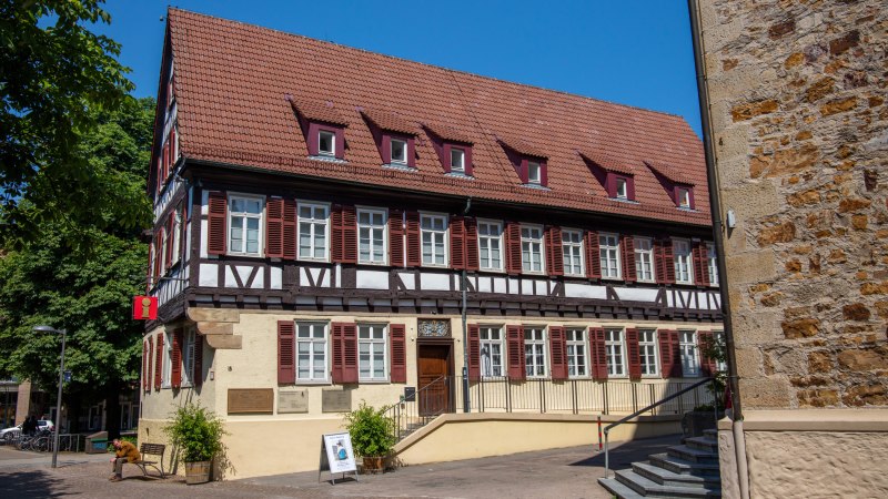 Fachwerkhaus mit rotem Dach und Tourist-Info-Schild. Vor dem Gebäude sitzt eine Person auf einer Bank. Blauer Himmel im Hintergrund., © Kirchheim unter Teck