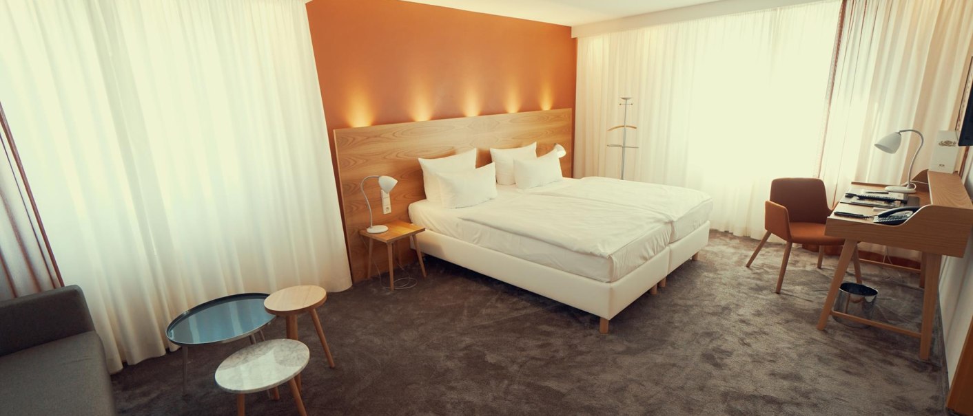 Helles Hotelzimmer mit Doppelbett, Schreibtisch, Sessel und kleinen Tischen. Große Fenster mit Vorhängen lassen viel Licht herein., © Ringhotel Adler Asperg