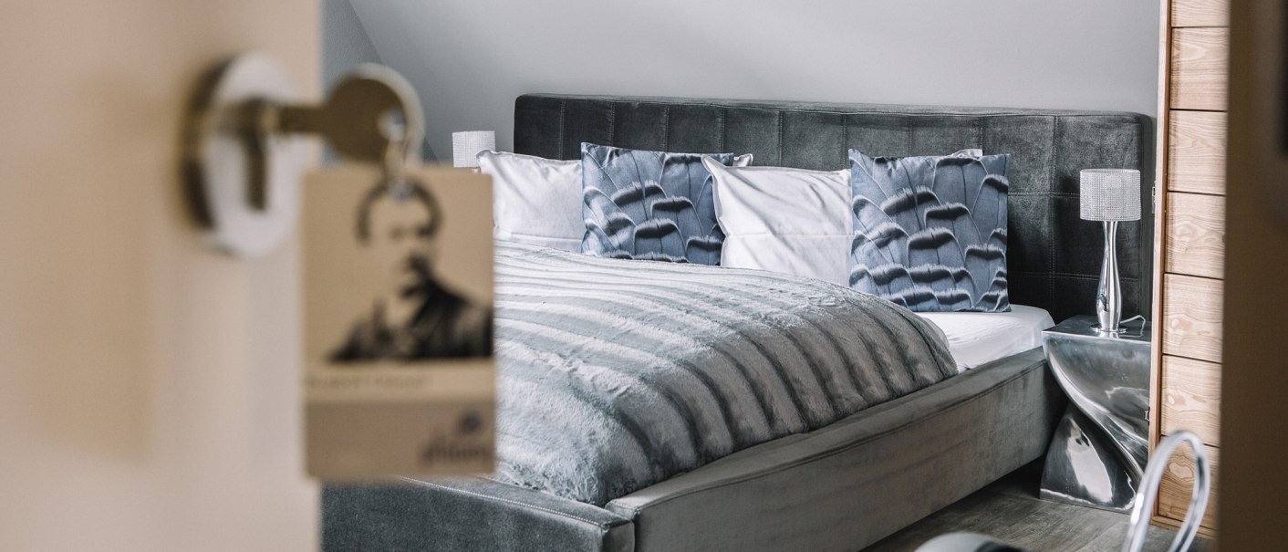 Ein elegantes Hotelzimmer mit einem großen, grauen Bett, dekorativen Kissen und einem modernen Nachttisch., © Boutiquehotel Pfauen Ein elegantes Hotelzimmer mit einem großen, grauen Bett, dekorativen Kissen und einem modernen Nachttisch., © Boutiquehotel Pfauen