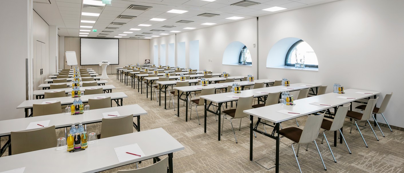 Moderner Tagungsraum mit wei&szlig;en Tischen und St&uuml;hlen, ausgestattet mit Getr&auml;nken und Schreibmaterialien. Ein Projektor und eine Leinwand sind vorhanden., &copy; ARCOTEL Hotels