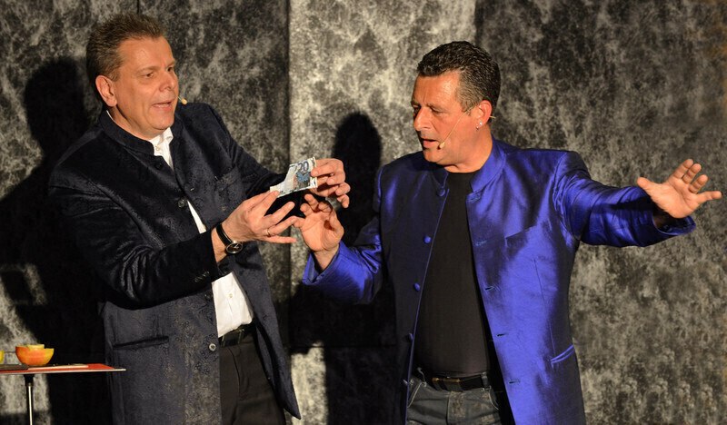 Zwei Männer in blauen Jacken führen auf einer Bühne einen Zaubertrick mit einem Geldschein vor., © LIMA-Theater Zwei Männer in blauen Jacken führen auf einer Bühne einen Zaubertrick mit einem Geldschein vor., © LIMA-Theater