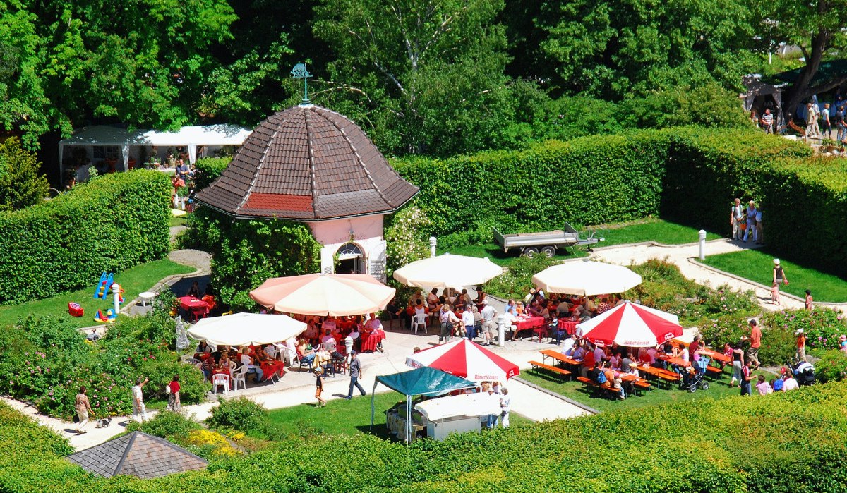 Ein Rosengarten mit Pavillon, umgeben von Hecken. Menschen sitzen unter bunten Sonnenschirmen und genießen das sonnige Wetter., © StaRT GmbH