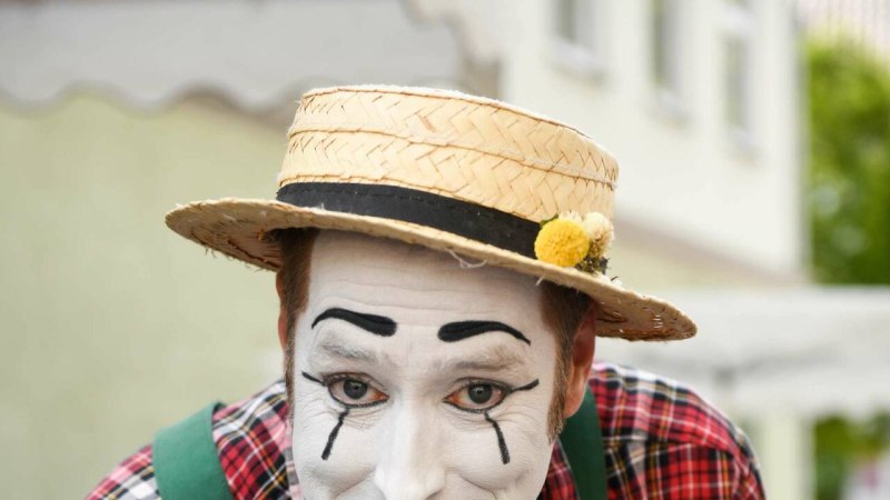 Ein Pantomime mit wei&szlig;em Gesicht und Strohhut zeigt auf eine Lupe mit Blume. Er tr&auml;gt ein kariertes Hemd und gr&uuml;ne Hosentr&auml;ger.