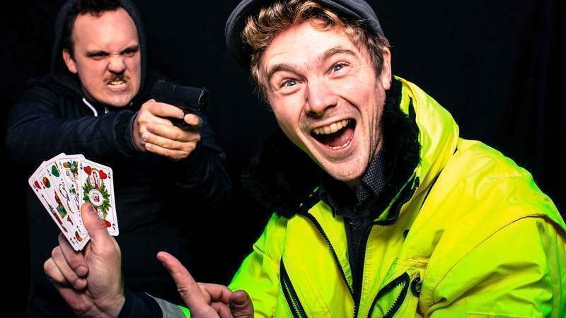 Ein Mann in gelber Jacke h&auml;lt lachend Spielkarten, w&auml;hrend ein anderer mit ernster Miene eine Spielzeugpistole auf ihn richtet., &copy; engesser marketing GmbH