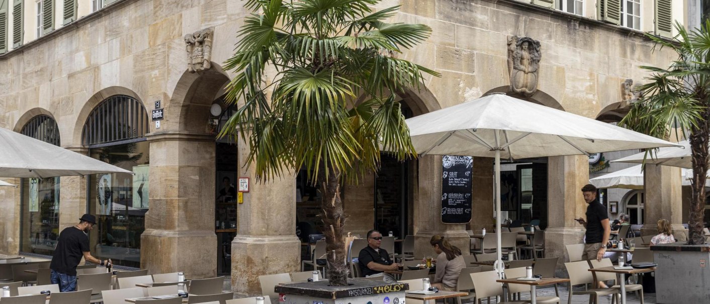 Café mit Tischen und Stühlen unter Sonnenschirmen vor einem historischen Gebäude mit Arkaden. Palmen und Menschen im Außenbereich., © SMG, Sarah Schmid