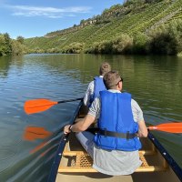 Zwei Personen paddeln in einem Kanu auf einem ruhigen Fluss, umgeben von grünen Bäumen und Weinbergen unter blauem Himmel., © Matthias Pflüger - Rock-the-River.com