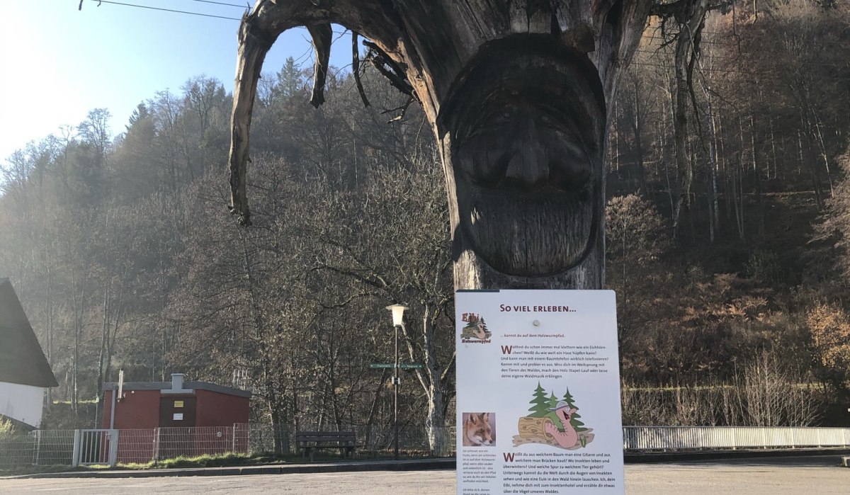 Ein geschnitzter Baumstamm mit Gesicht und Infotafel steht vor einer Straße, umgeben von bewaldeten Hügeln., © Nördlicher Schwarzwald