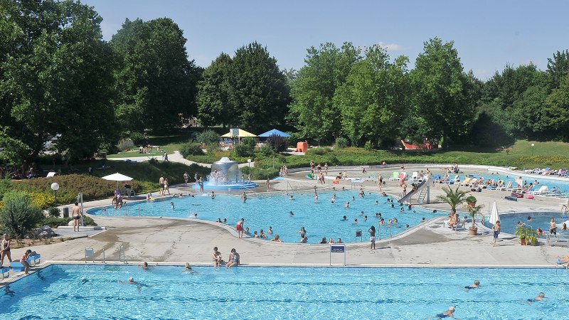 Ein belebtes Freibad mit mehreren Pools, vielen Besuchern und umgeben von gr&uuml;nen B&auml;umen. Menschen schwimmen und entspannen auf Liegest&uuml;hlen., &copy; Werner Kuhnle