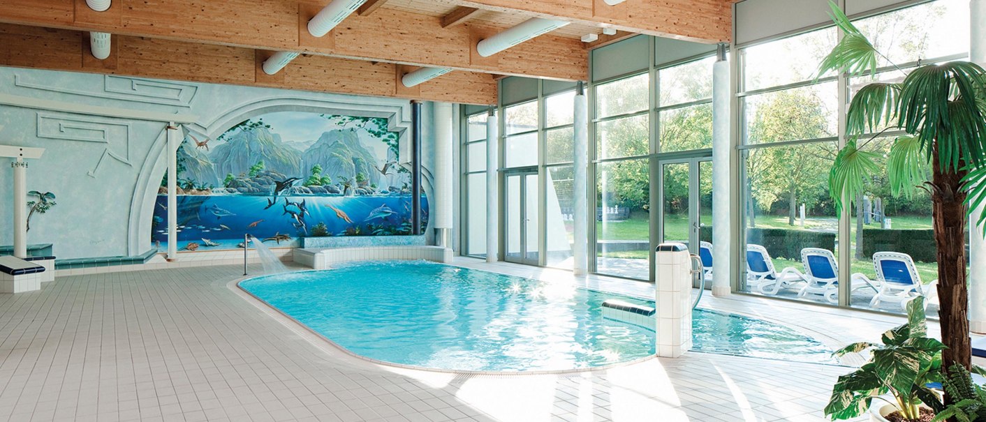Ein Innenpool mit einem Wandgemälde von Meerestieren. Große Fenster lassen viel Licht herein. Liegestühle stehen am Rand, eine Palme im Vordergrund., © Seminaris Hotel Bad Boll Ein Innenpool mit einem Wandgemälde von Meerestieren. Große Fenster lassen viel Licht herein. Liegestühle stehen am Rand, eine Palme im Vordergrund., © Seminaris Hotel Bad Boll