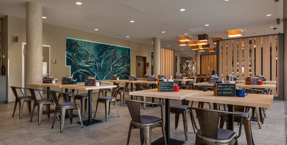 Moderner Frühstücksraum mit Holztischen, Metallstühlen und einem großen Wandbild. Die Beleuchtung ist warm und einladend., © Novum Hospitality Moderner Frühstücksraum mit Holztischen, Metallstühlen und einem großen Wandbild. Die Beleuchtung ist warm und einladend., © Novum Hospitality