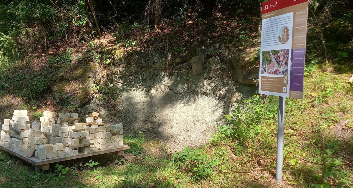 Geopfad Station 13 in Gaildorf: Eine Infotafel steht neben einem Haufen heller Steine auf einer Holzpalette, umgeben von Wald. Geopfad Station 13 in Gaildorf: Eine Infotafel steht neben einem Haufen heller Steine auf einer Holzpalette, umgeben von Wald.