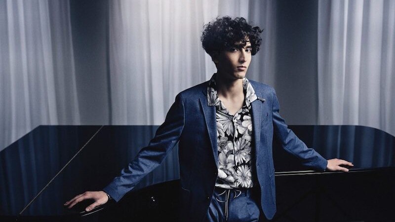 Ein Mann in einem blauen Anzug mit gemustertem Hemd steht vor einem Flügel. Der Hintergrund ist mit weißen Vorhängen gestaltet., © Kammerkonzerte Schwäbisch Hall e.V.