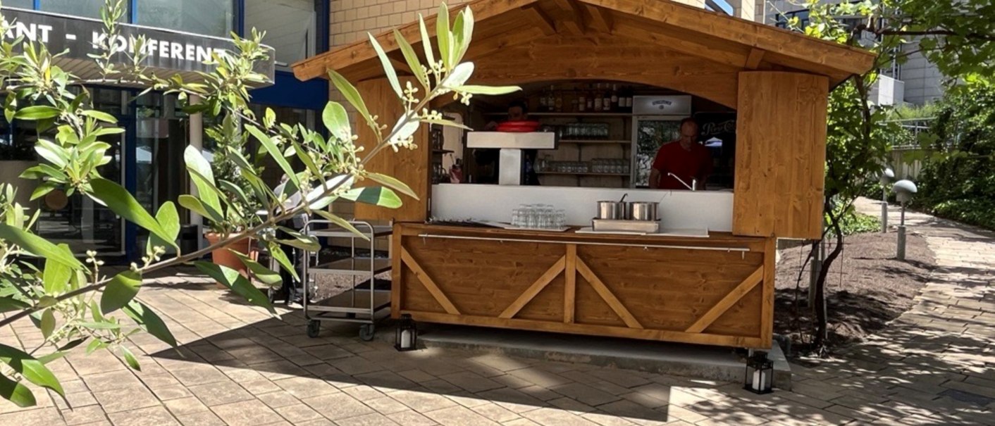 Ein Holzstand vor einem modernen Gebäude mit Glasfassade. Im Vordergrund sind Pflanzen zu sehen, die den Stand teilweise verdecken., © TOMAS Ein Holzstand vor einem modernen Gebäude mit Glasfassade. Im Vordergrund sind Pflanzen zu sehen, die den Stand teilweise verdecken., © TOMAS