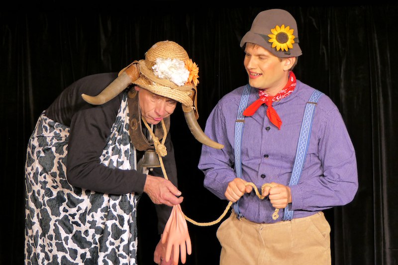 Zwei Schauspieler in einem humorvollen Theaterst&uuml;ck. Einer tr&auml;gt ein Kuhkost&uuml;m, der andere einen Hut mit Sonnenblume. Beide scheinen in eine Szene vertieft zu sein., &copy; Galli Theater Backnang