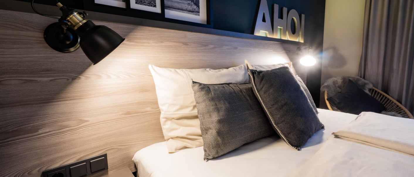 Modernes Schlafzimmer mit einem Bett, grauen Kissen, Wandlampen und Dekorationen wie Bilderrahmen und dem Schriftzug 'AHOI' an der Wand., © HARBR. hotel Ludwigsburg / DQuadrat Living GmbH Modernes Schlafzimmer mit einem Bett, grauen Kissen, Wandlampen und Dekorationen wie Bilderrahmen und dem Schriftzug 'AHOI' an der Wand., © HARBR. hotel Ludwigsburg / DQuadrat Living GmbH