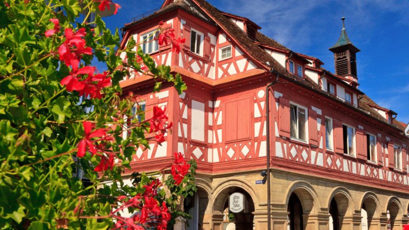 Das Alte Rathaus in Waiblingen, ein Fachwerkgeb&auml;ude mit roten und wei&szlig;en Details, umgeben von bl&uuml;henden roten Blumen unter blauem Himmel., &copy; WTM GmbH, Kai Koepf