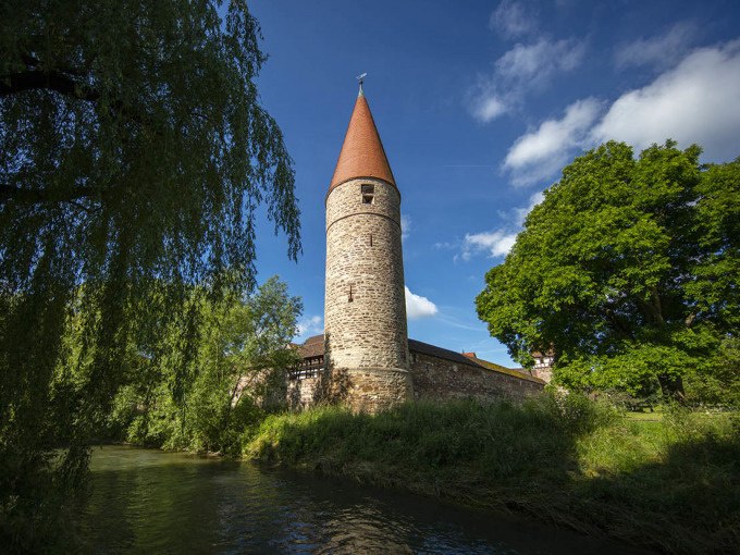 Ein mittelalterlicher Turm mit rotem Dach steht malerisch neben einem Fluss, umgeben von &uuml;ppigem Gr&uuml;n und einer alten Steinmauer., &copy; Stadtverwaltung Weil der Stadt