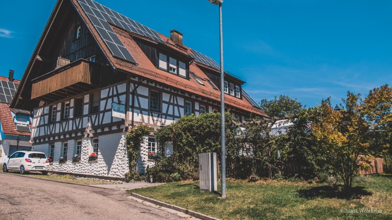 Fachwerkhaus mit Solarpanelen auf dem Dach, umgeben von Bäumen und einem Auto. Klare blaue Himmel und eine Straßenlaterne im Vordergrund., © Stadt Welzheim