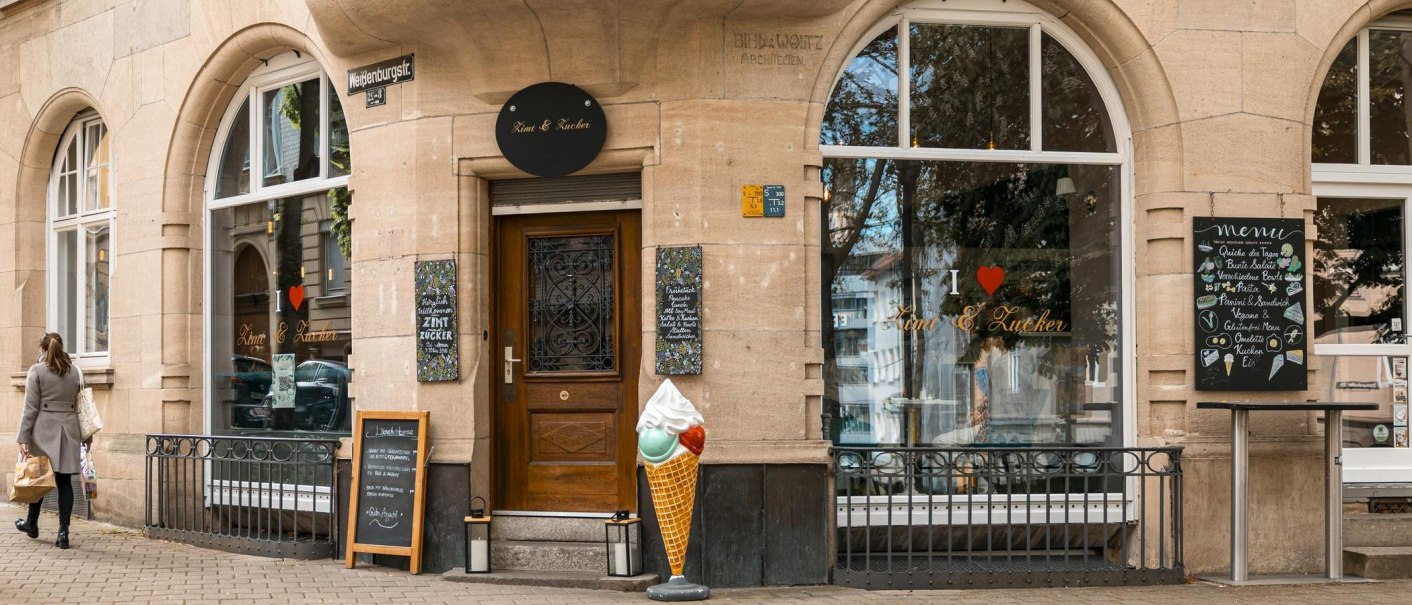 Ein Café namens 'Zimt & Zucker' an einer Straßenecke mit großen Fenstern, einem Eistütenmodell und einer Frau, die vorbeigeht., © SMG, Sarah Schmid Ein Café namens 'Zimt & Zucker' an einer Straßenecke mit großen Fenstern, einem Eistütenmodell und einer Frau, die vorbeigeht., © SMG, Sarah Schmid