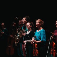 Musiker auf einer Bühne mit Hörnern, Geigen und Flöten, in dunkler Umgebung, lächelnd und konzentriert., © Sky Bürhaus