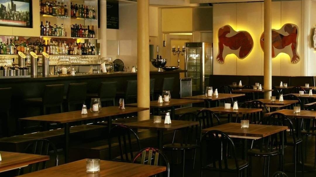 Leeres Restaurant mit Bar, Holztischen und schwarzen Stühlen. An der Wand hängen Kunstwerke. Die Beleuchtung ist warm und einladend., © Blauer Engel GmbH