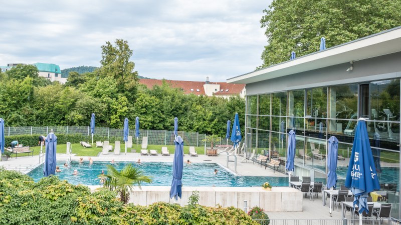 Ein Reha-Zentrum mit Pool, Liegest&uuml;hlen und blauen Sonnenschirmen. Im Hintergrund sind B&auml;ume und Geb&auml;ude zu sehen., &copy; Christophsbad Klinikgruppe