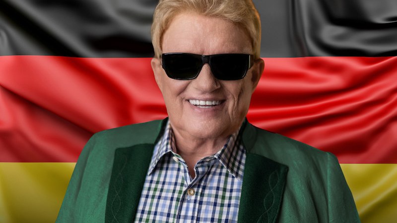 Eine Person mit Sonnenbrille und kariertem Hemd steht vor einer deutschen Flagge. Sie trägt einen grünen Blazer und lächelt in die Kamera., © Thomann Künstlermanagement GmbH Eine Person mit Sonnenbrille und kariertem Hemd steht vor einer deutschen Flagge. Sie trägt einen grünen Blazer und lächelt in die Kamera., © Thomann Künstlermanagement GmbH