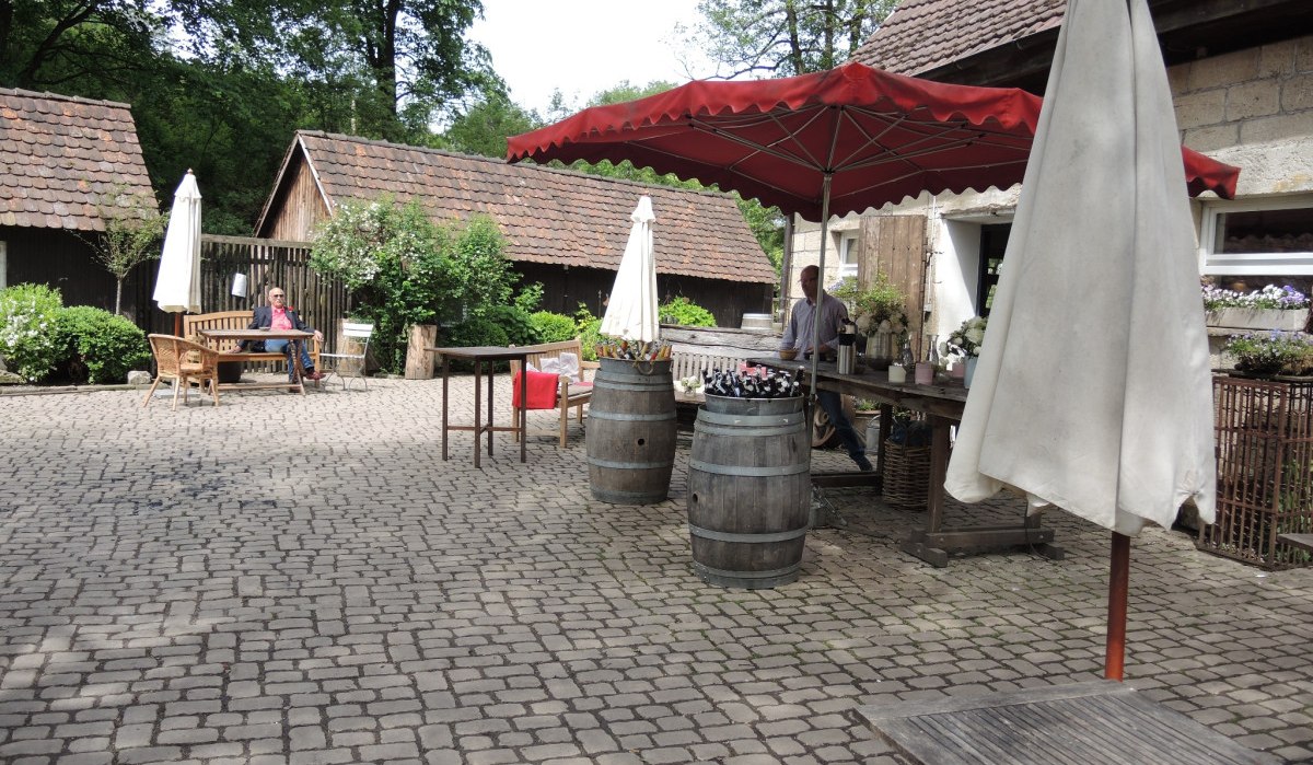 Ein gepflasterter Hof mit Tischen, Stühlen und Sonnenschirmen vor alten Gebäuden. Eine Person sitzt auf einer Bank., © Natur.Nah. Schönbuch & Heckengäu Ein gepflasterter Hof mit Tischen, Stühlen und Sonnenschirmen vor alten Gebäuden. Eine Person sitzt auf einer Bank., © Natur.Nah. Schönbuch & Heckengäu