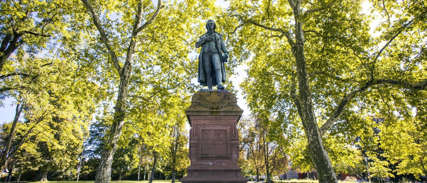 Das Schiller-Denkmal in Marbach am Neckar steht majestätisch zwischen grünen Bäumen, die im Sonnenlicht leuchten., © SMG, Sarah Schmid Das Schiller-Denkmal in Marbach am Neckar steht majestätisch zwischen grünen Bäumen, die im Sonnenlicht leuchten., © SMG, Sarah Schmid