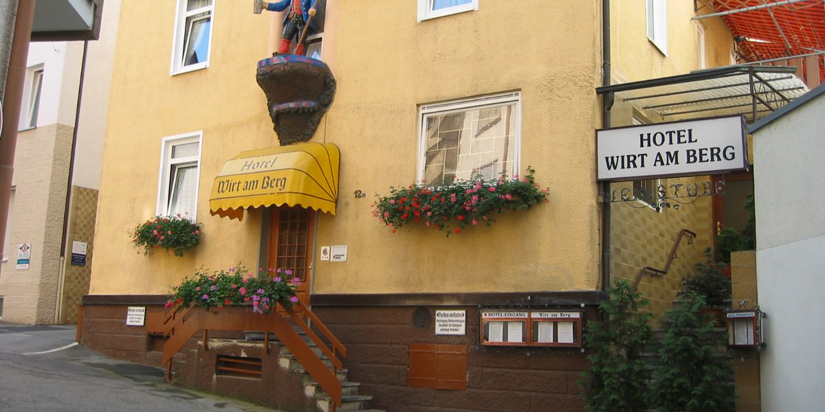 Gelbes Gebäude mit dem Schild 'Hotel Wirt am Berg'. Über dem Eingang eine Figur, Blumenkästen an den Fenstern. Rechts ein weiteres Schild mit Hotelnamen., © Hotel Wirt am Berg
