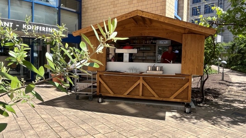 Ein Holzstand vor einem modernen Gebäude mit Glasfassade. Im Vordergrund sind Pflanzen zu sehen, die den Stand teilweise verdecken., © TOMAS Ein Holzstand vor einem modernen Gebäude mit Glasfassade. Im Vordergrund sind Pflanzen zu sehen, die den Stand teilweise verdecken., © TOMAS