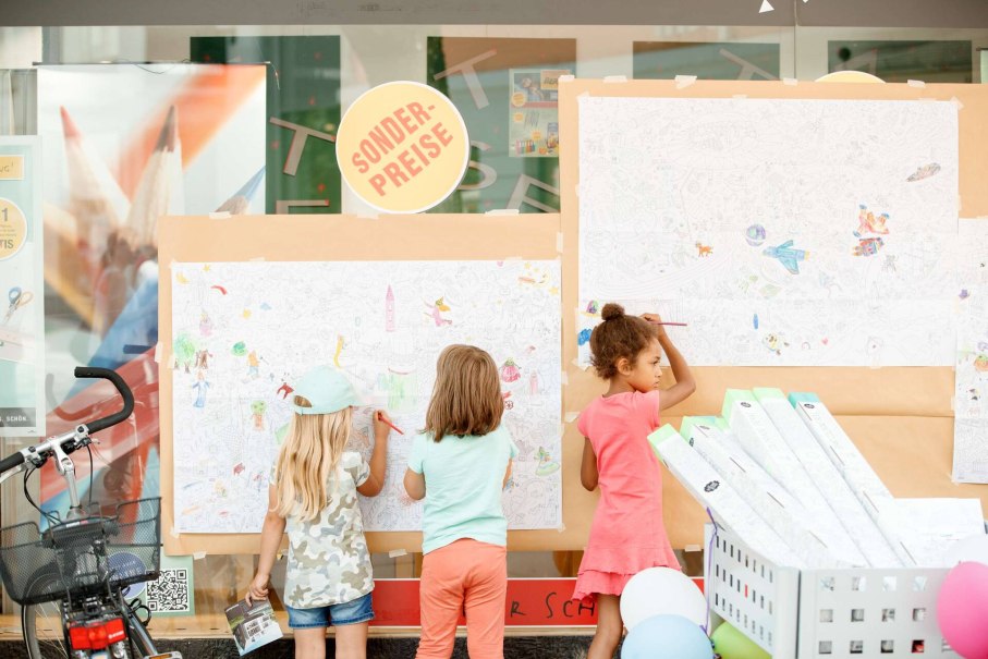 Drei Kinder malen an gro&szlig;en Ausmalpostern vor einem Schaufenster. Ein Sonderpreis-Schild ist sichtbar. Ein Fahrrad steht daneben.