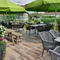 Gem&uuml;tliche Terrasse eines Landgasthofs mit gr&uuml;nen Sonnenschirmen, Holztischen und St&uuml;hlen. Blumen schm&uuml;cken den Bereich, im Hintergrund Fachwerkh&auml;user., &copy; Landgasthof Linde