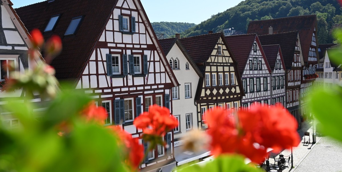 Fachwerkhäuser am Marktplatz von Bad Urach, umgeben von grünen Hügeln. Rote Blumen im Vordergrund verleihen dem Bild Farbe., © Bad Urach Tourismus Fachwerkhäuser am Marktplatz von Bad Urach, umgeben von grünen Hügeln. Rote Blumen im Vordergrund verleihen dem Bild Farbe., © Bad Urach Tourismus