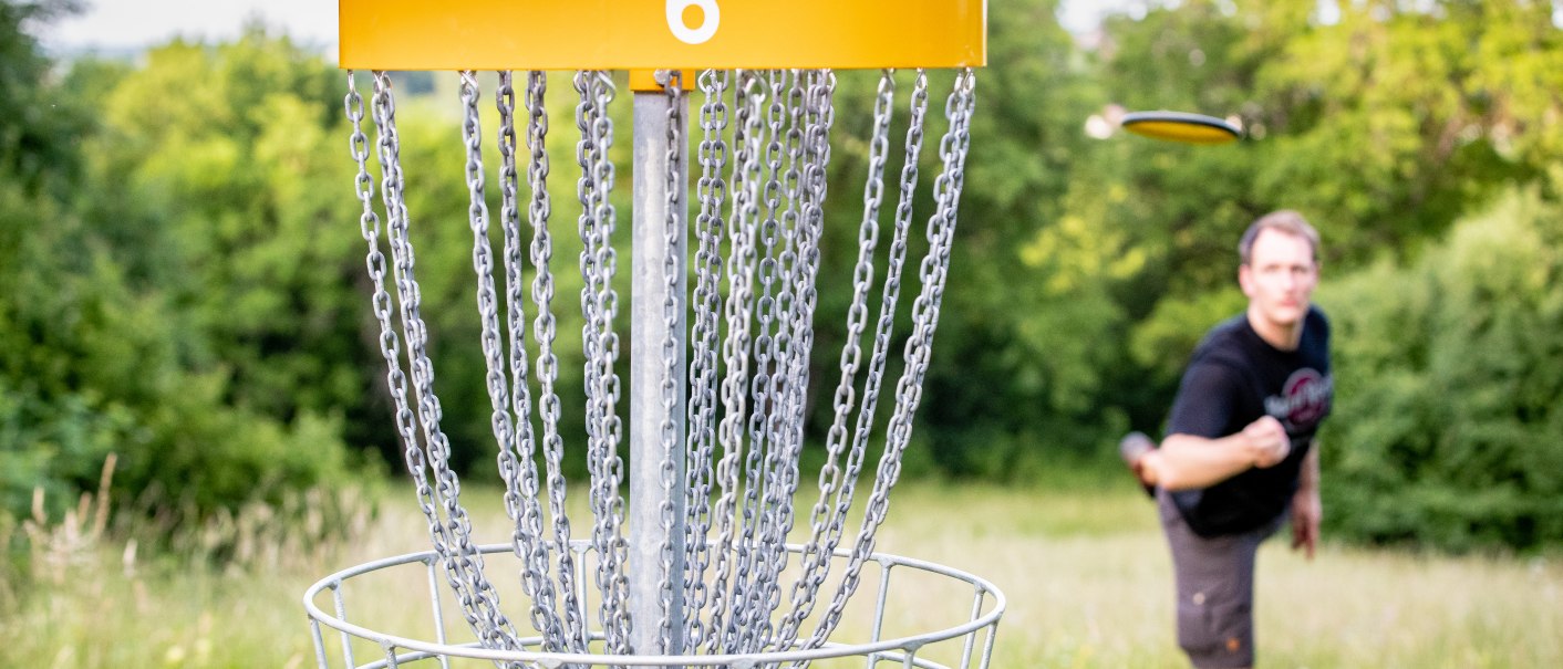 Ein Discgolf-Korb mit der Nummer 6 im Vordergrund, ein Spieler wirft eine Scheibe im Hintergrund auf einer grünen Wiese., © Benjamin Lau