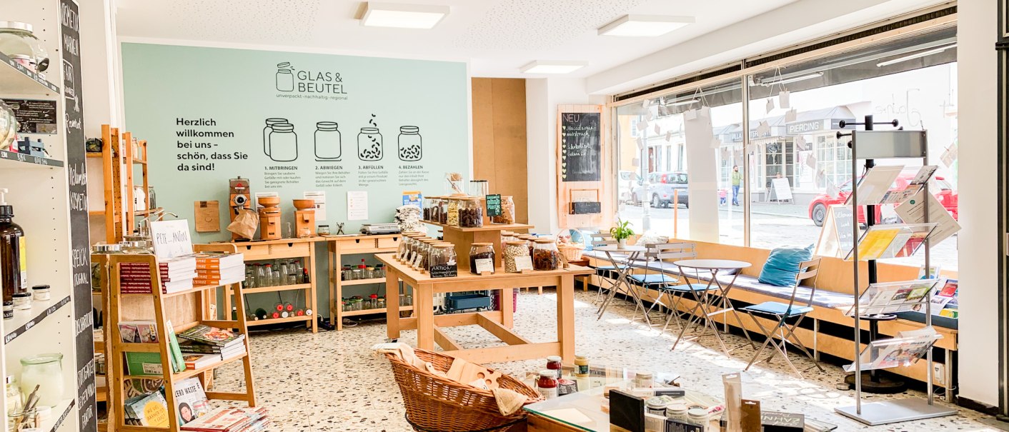 Ein heller Unverpackt-Laden mit Regalen voller Produkte, Tischen und einer gro&szlig;en Fensterfront. An der Wand steht "Glas & Beutel"., &copy; cGlasundBeutel